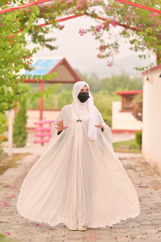 Woman wearing a flowing white abaya and hijab with black face mask outdoors under flowering trees