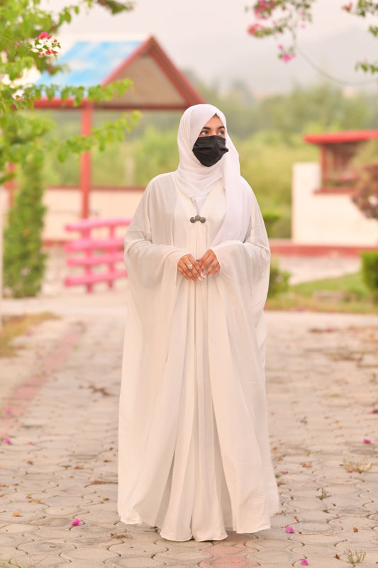 Woman wearing a white abaya and hijab with a black face mask standing outdoors in a garden setting