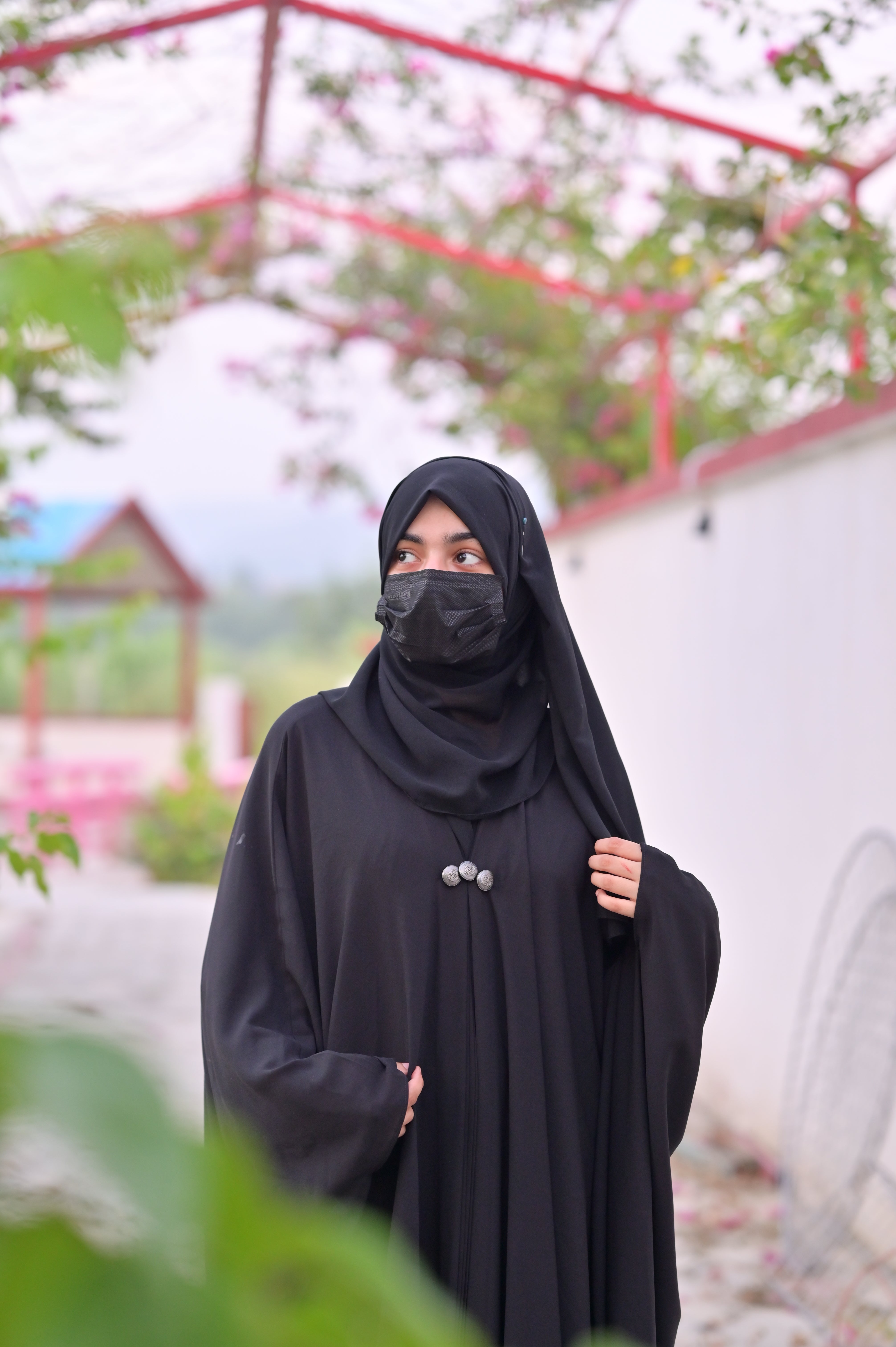 Woman wearing a black abaya with matching scarf and face mask standing outdoors near a garden