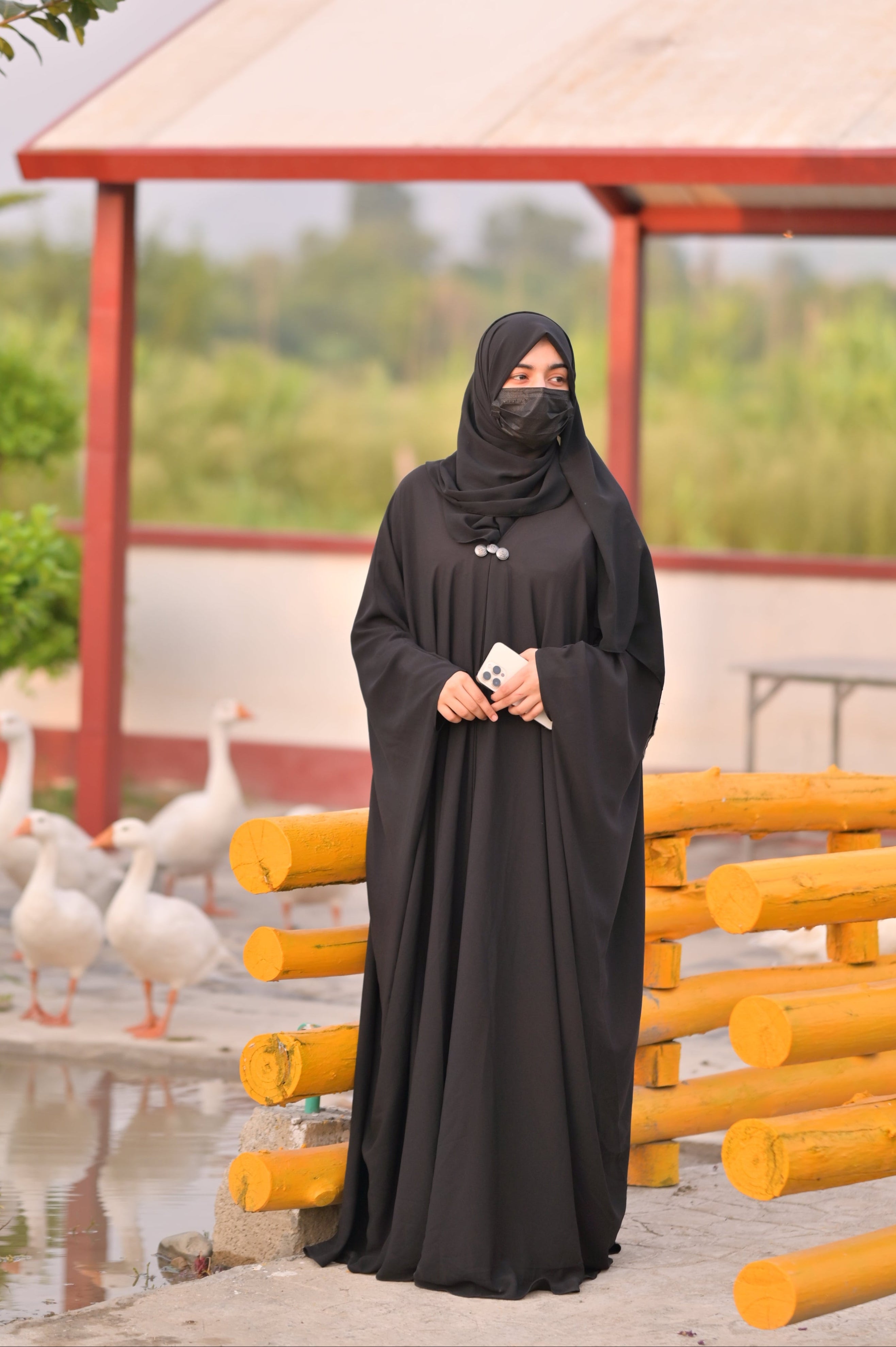 Black abaya with matching scarf worn by a woman standing outdoors near a wooden fence and white ducks