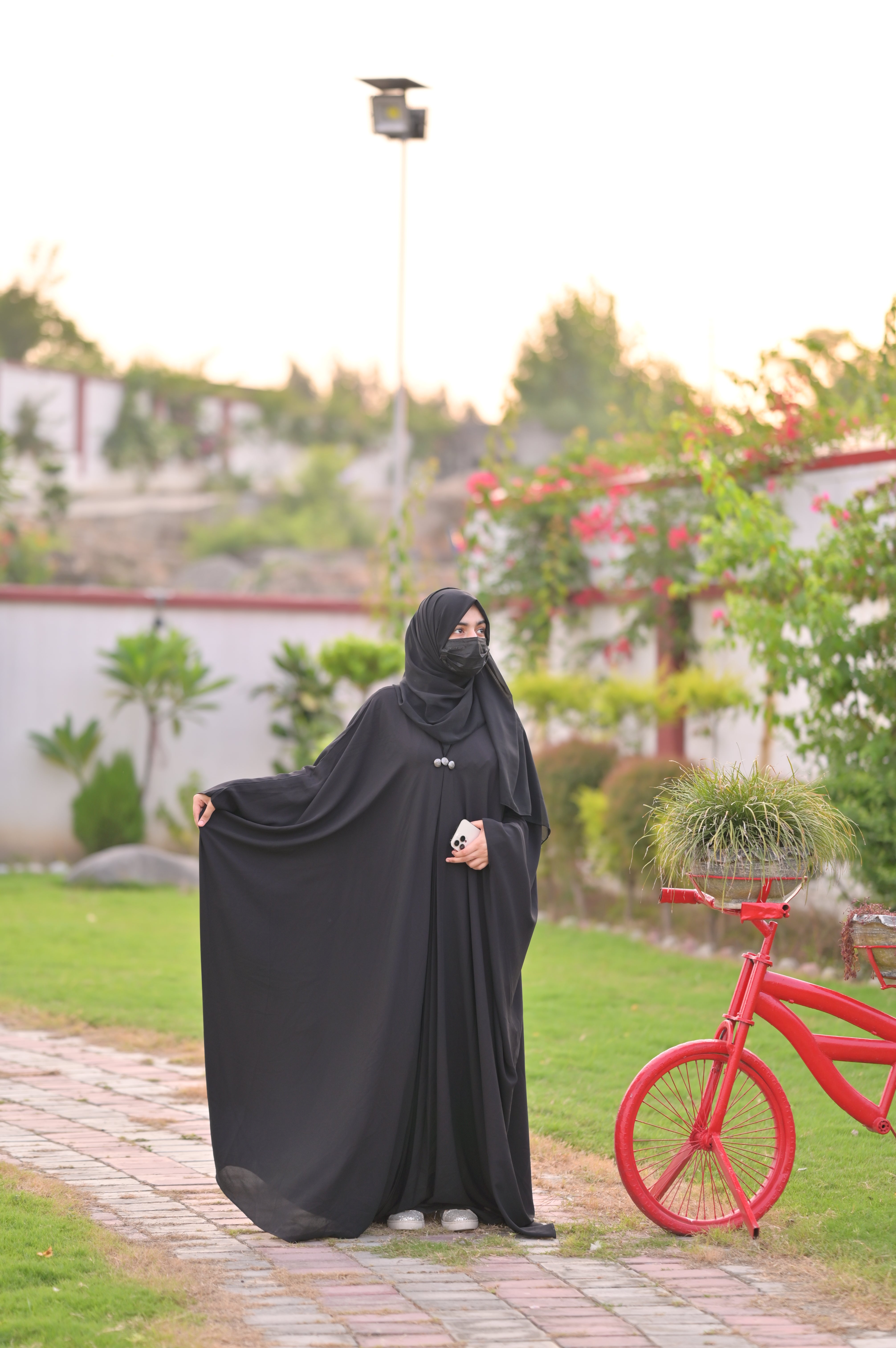 Woman wearing black abaya with matching scarf standing outdoors near a red bicycle in a garden