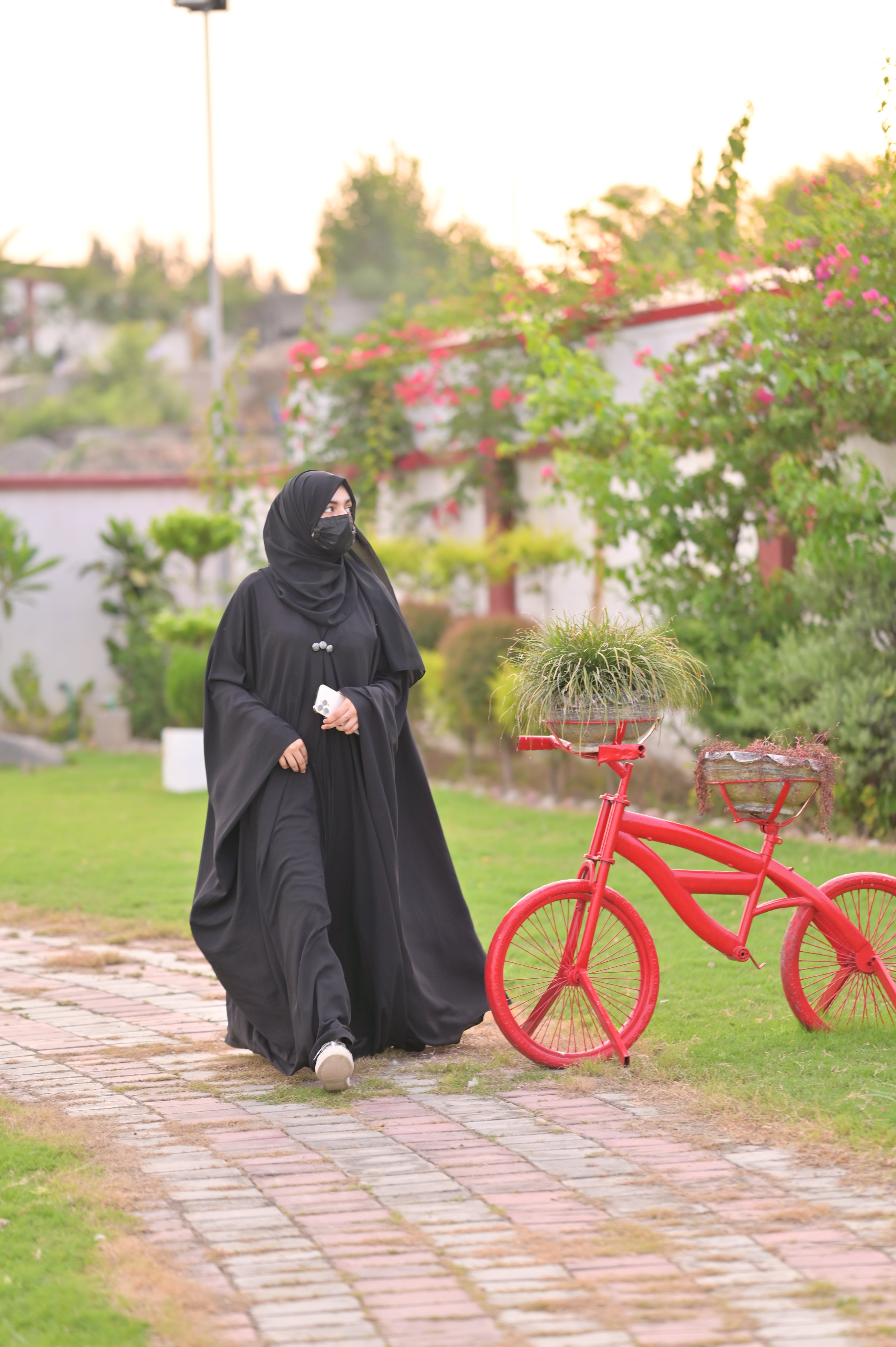 Woman wearing black abaya with matching scarf walking on garden path near red bicycle
