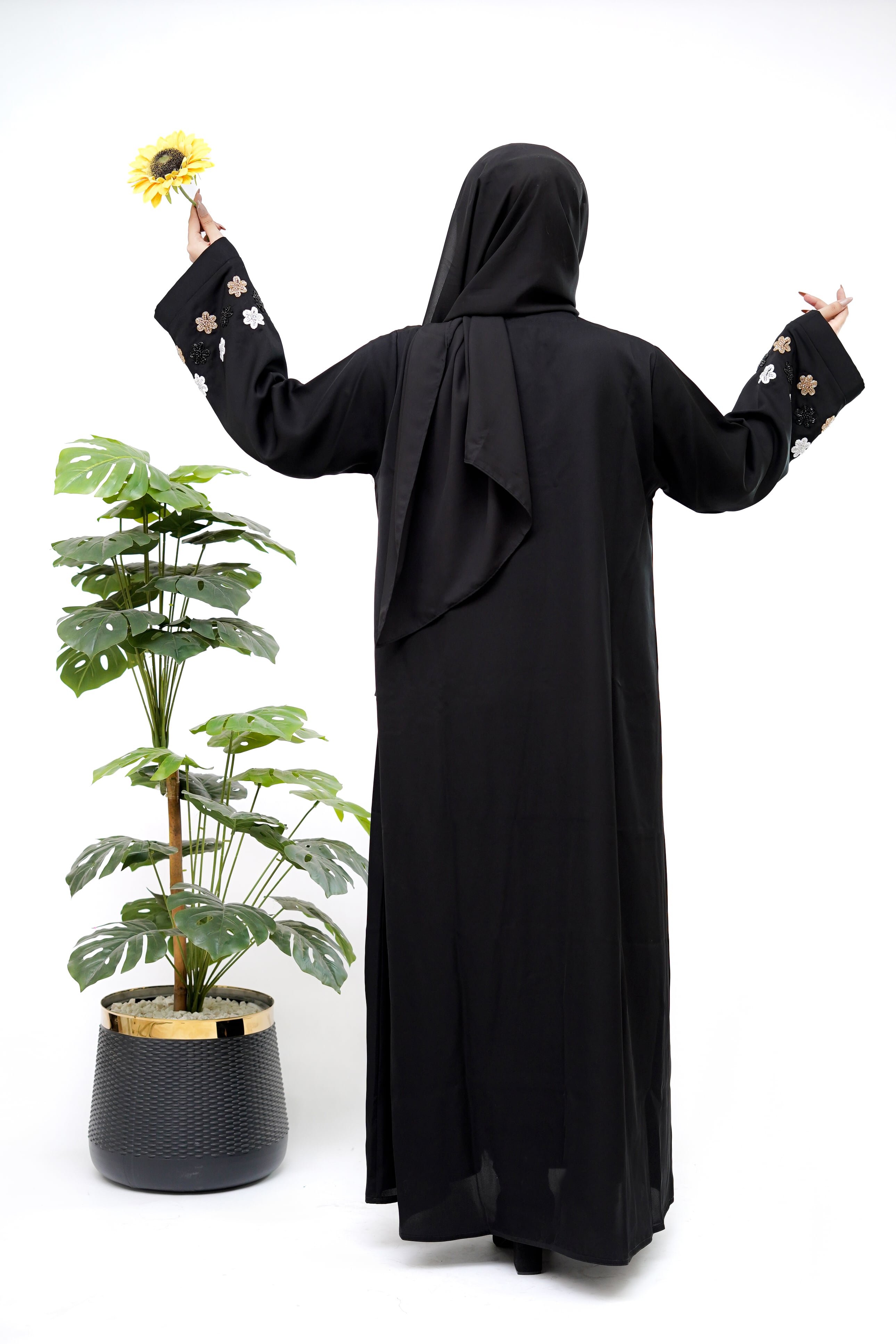 Woman wearing the Best Black Abaya from Clarity Glam with floral embroidered sleeves posing next to a green potted plant