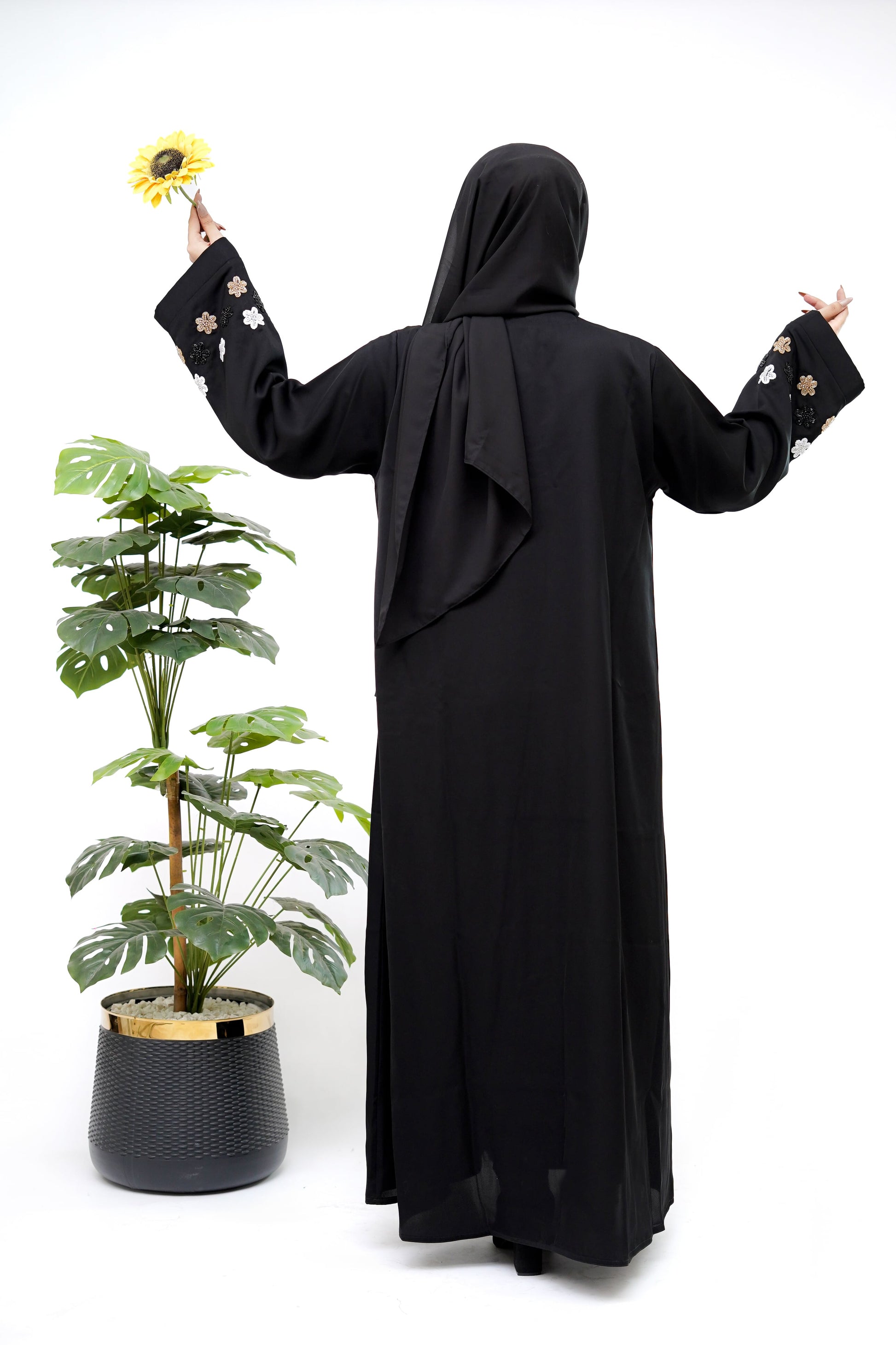 Woman wearing the Best Black Abaya from Clarity Glam with floral embroidered sleeves posing next to a green potted plant