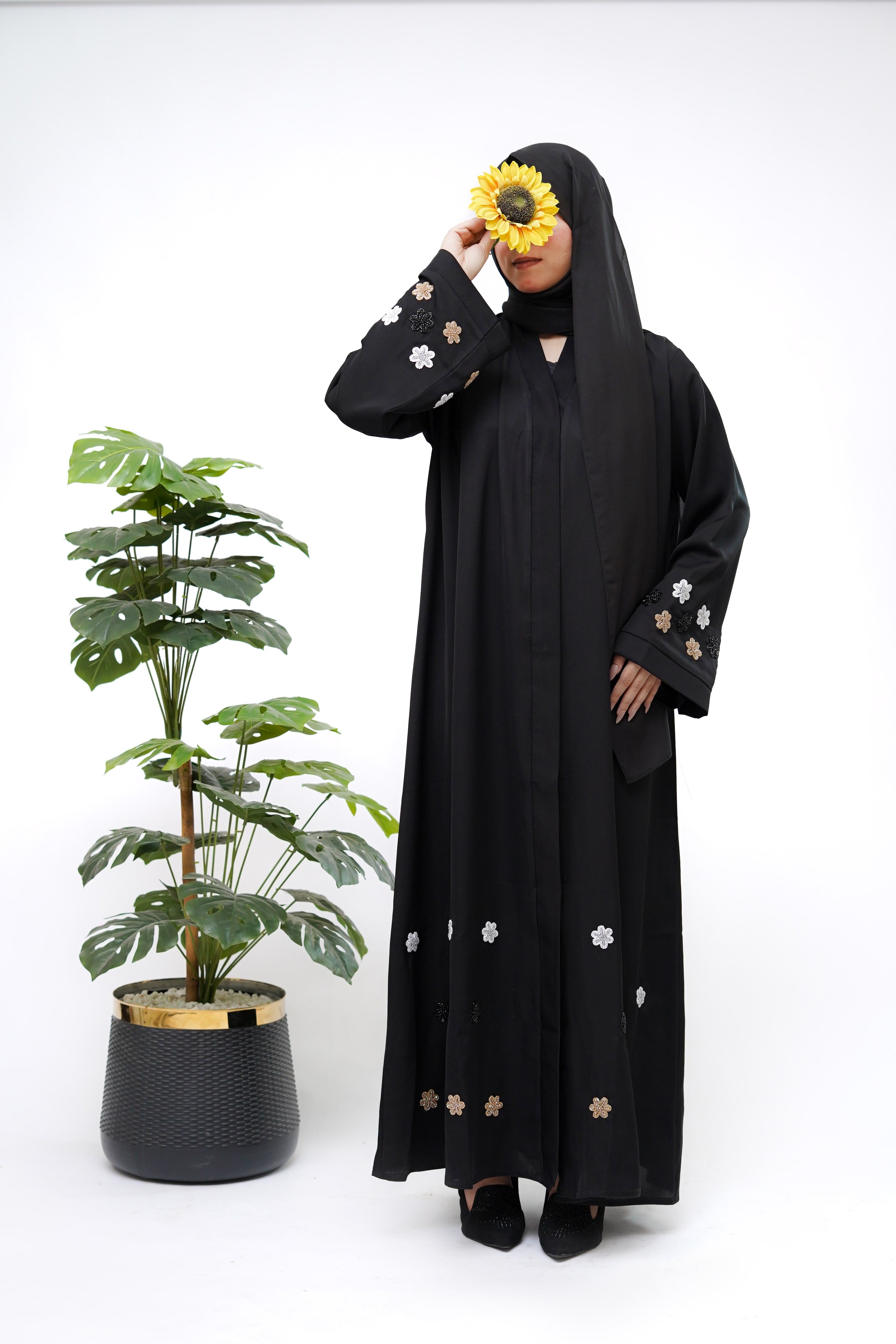 Woman wearing best black abaya with floral embroidery standing next to a potted plant