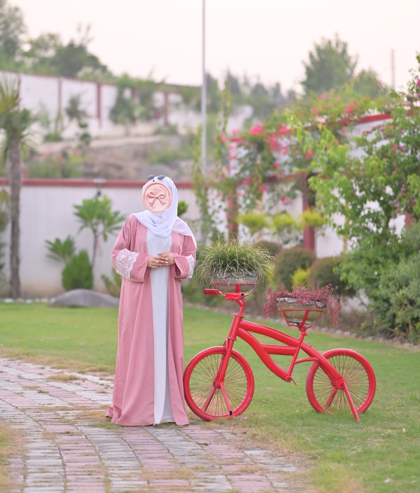 Clarity Glam luxury abaya with scarf in soft pink worn outdoors near red bicycle and greenery