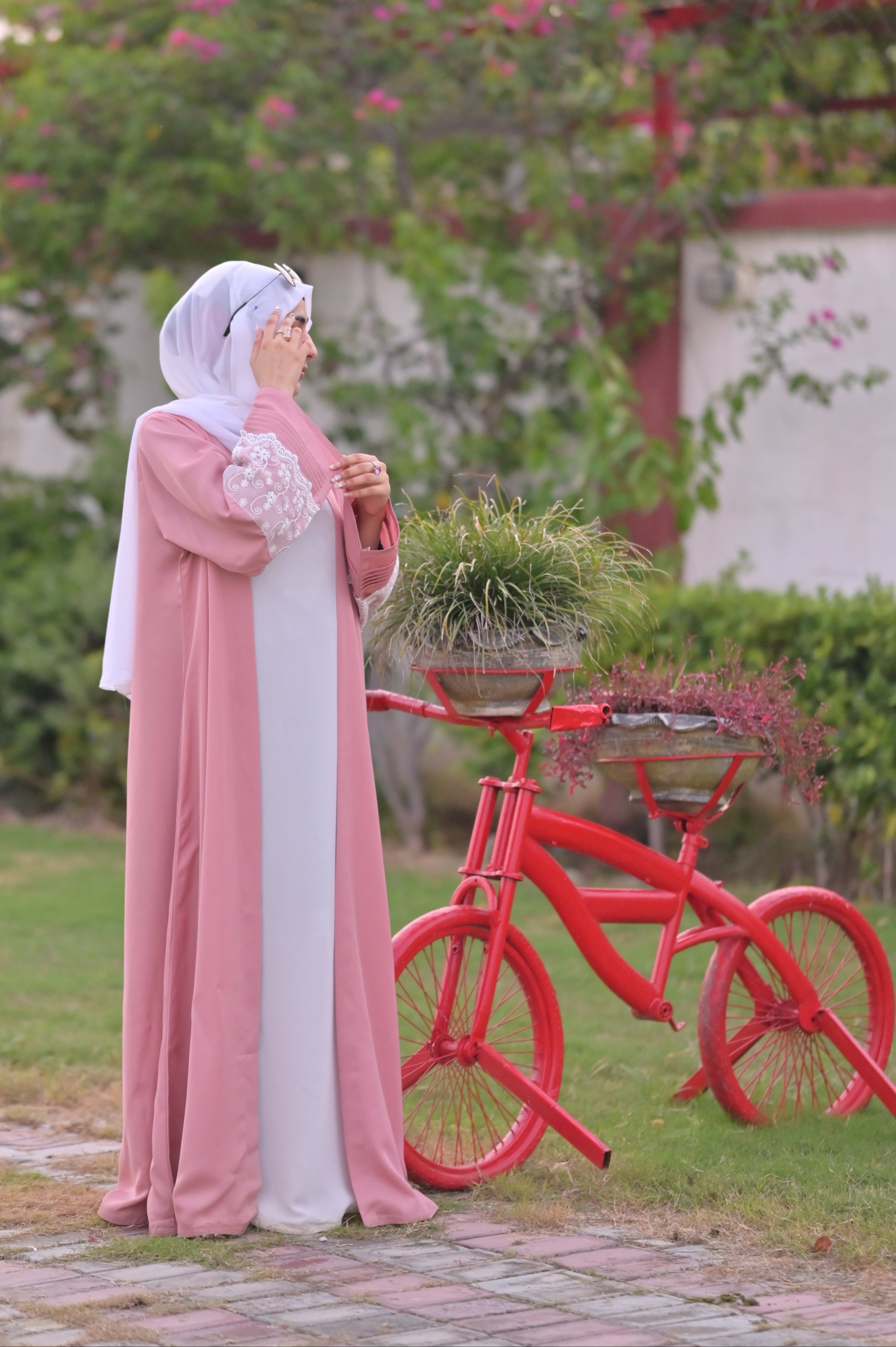 Clarity Glam luxury abaya with scarf in pink and white party wear standing next to decorative red bicycle planter