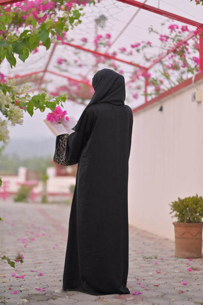 Woman wearing luxury black abaya with intricate sleeve details standing outdoors under flowering trellis
