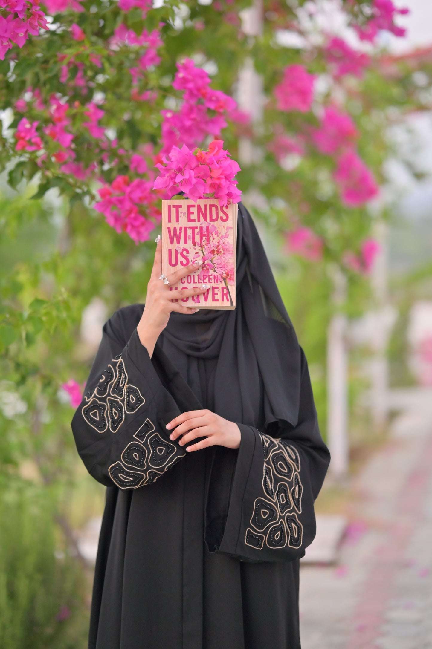 Woman wearing a luxury black abaya with intricate sleeve designs holding a book in front of pink flowers