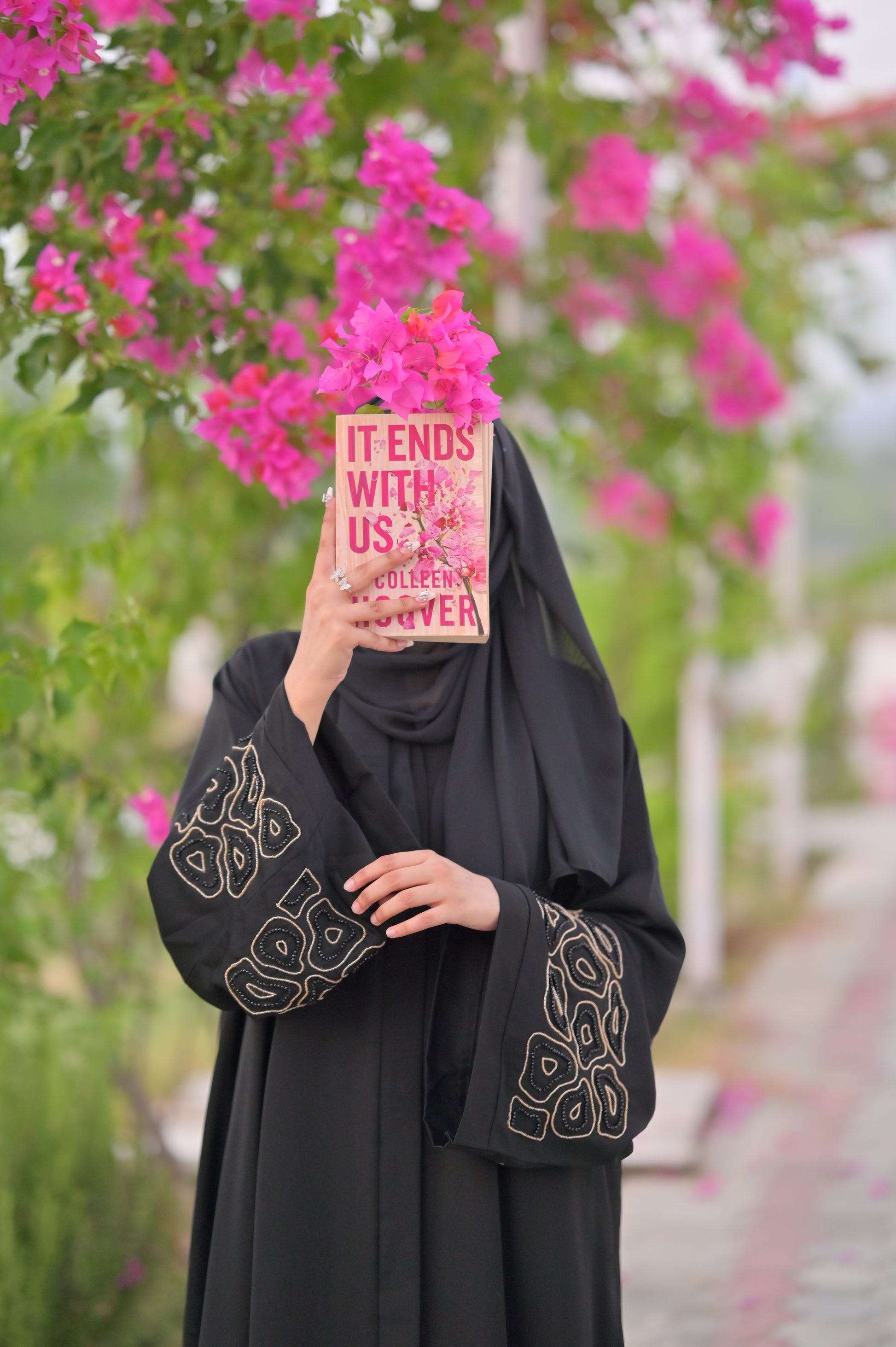 Woman wearing a luxury black abaya with intricate sleeve designs holding a book in front of pink flowers