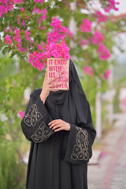Woman wearing a luxury black abaya with intricate sleeve designs holding a book in front of pink flowers