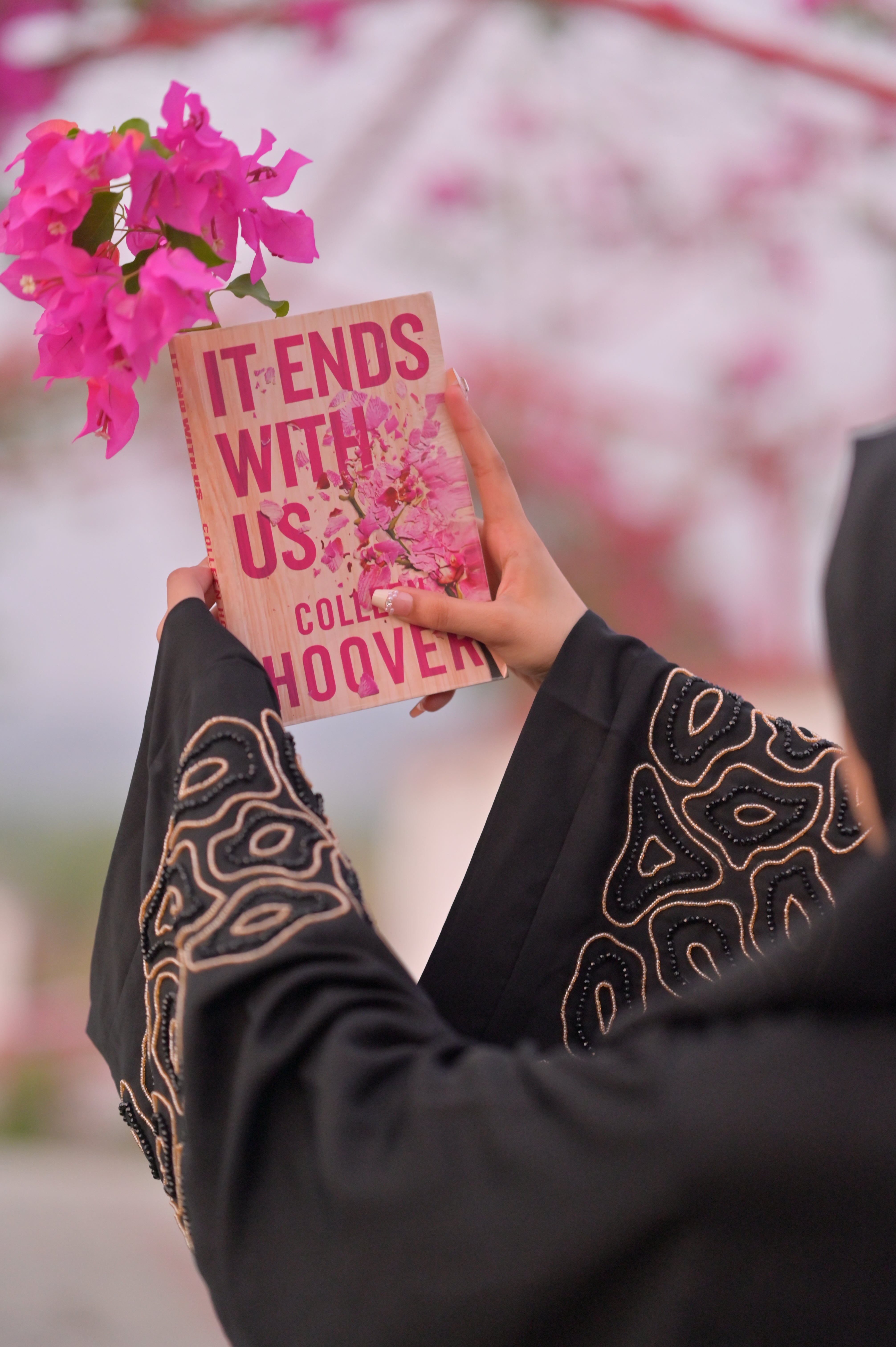 Woman wearing luxury black abaya with gold embroidery holding a book and pink flowers outdoors