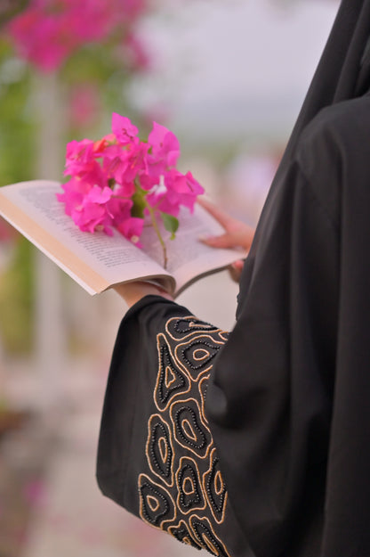 Clarity Glam luxury black abaya with intricate gold embroidery on sleeve holding a book and pink flowers